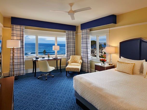 offers-ocean-view-deal A hotel room with a bed, dresser, tv, chairs and a desk arranged in front of two windows with a view of the ocean